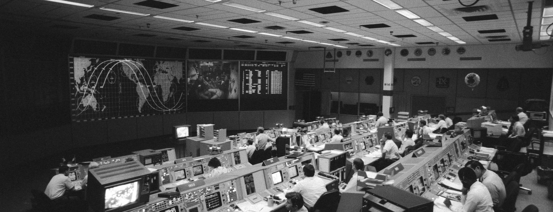 A wide view of a NASA flight control room, rows of consoles facing a projected world map.
